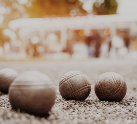 Close-up of four metallic bocce balls on a gravel ground, with a blurred outdoor background showing people and trees.